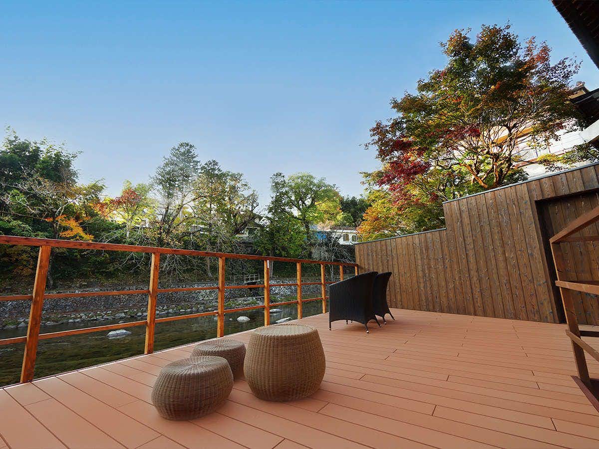 Ikedaya Kajikaen Wooden deck platform with horizontal-bar railing, woven seat cushions, trees with foliage in background, landscape area, clear sky.