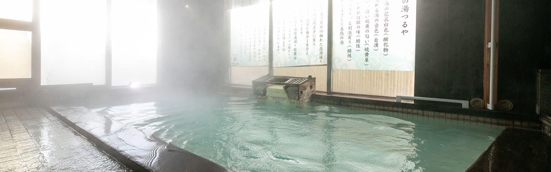 Gokan no Yu Tsuruy indoor bath with turquoise water, white tile floor, left windows with daylight, and signage on dark wall