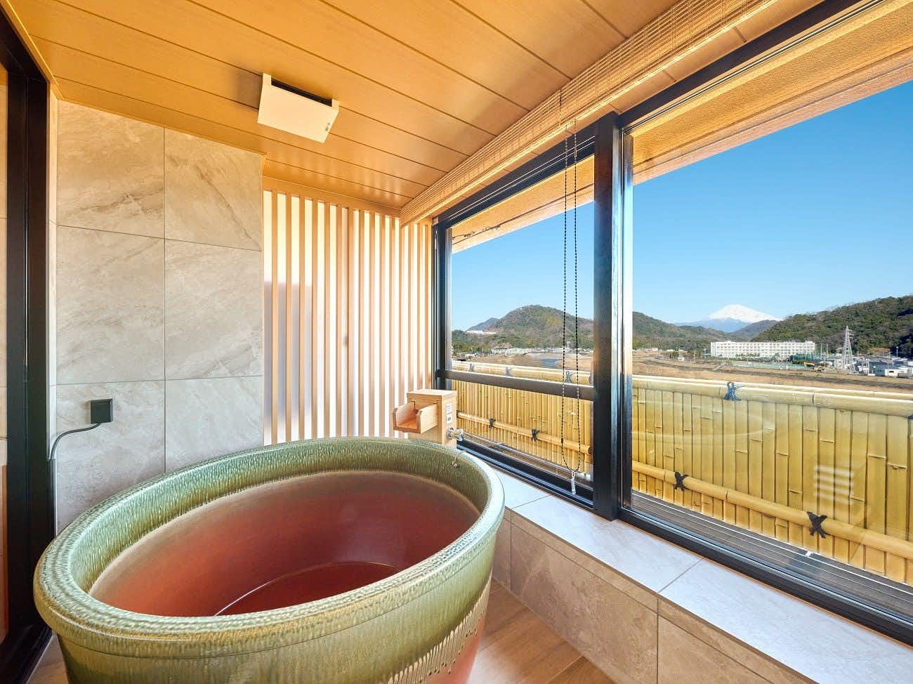 Fugaku Hanabusa Bath with pale rim and reddish water, wooden ceiling, marble walls, and large window overlooking mountain valley.