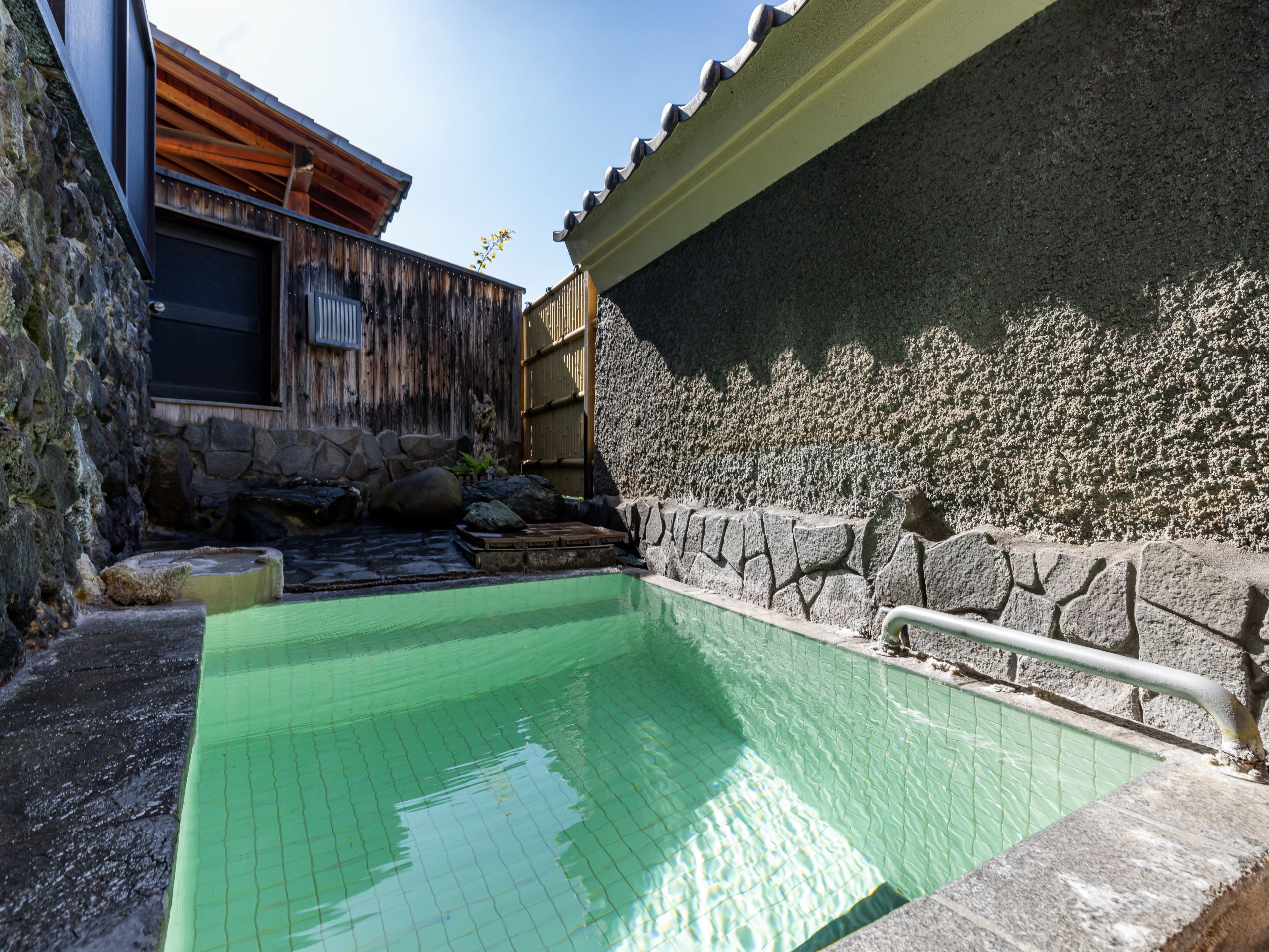 A turquoise pool runs beside a dark stone wall and wooden building.