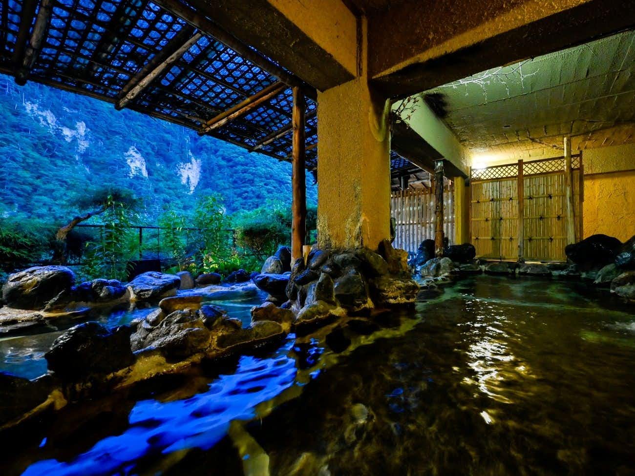 Gorokaku Roofed rock-lined bath under evening light with timber posts, bamboo screening, and a wooded hillside beyond