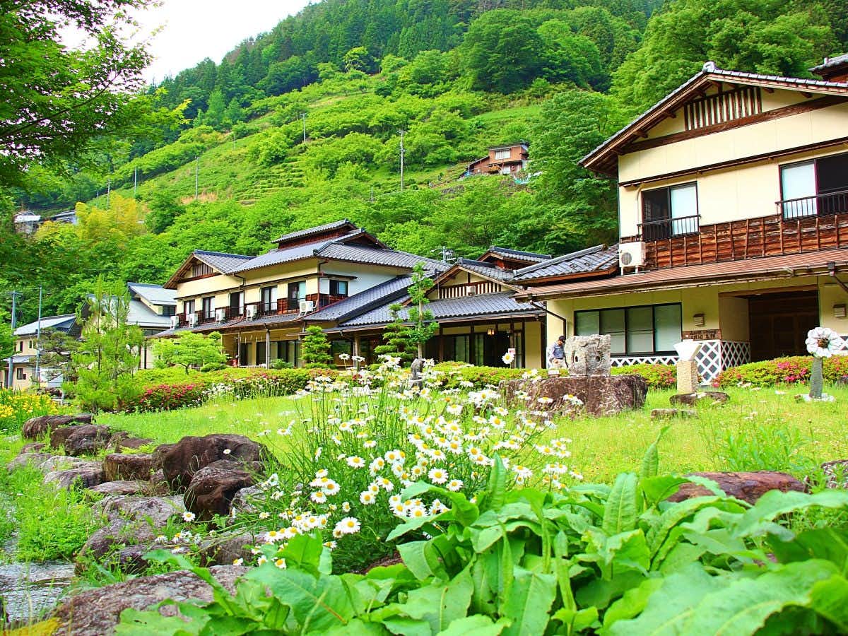 Irori no Yado Shimabatake with this scene, where a quiet mountain hamlet spreads across green slopes beyond flowering fields.