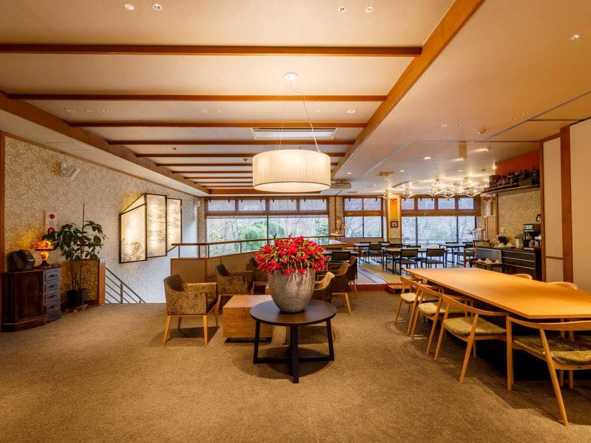 Kokoro no Oyado Jizai-so Spacious lounge and dining area with long wood tables, upholstered chairs, beams, broad windows, and bright flowers