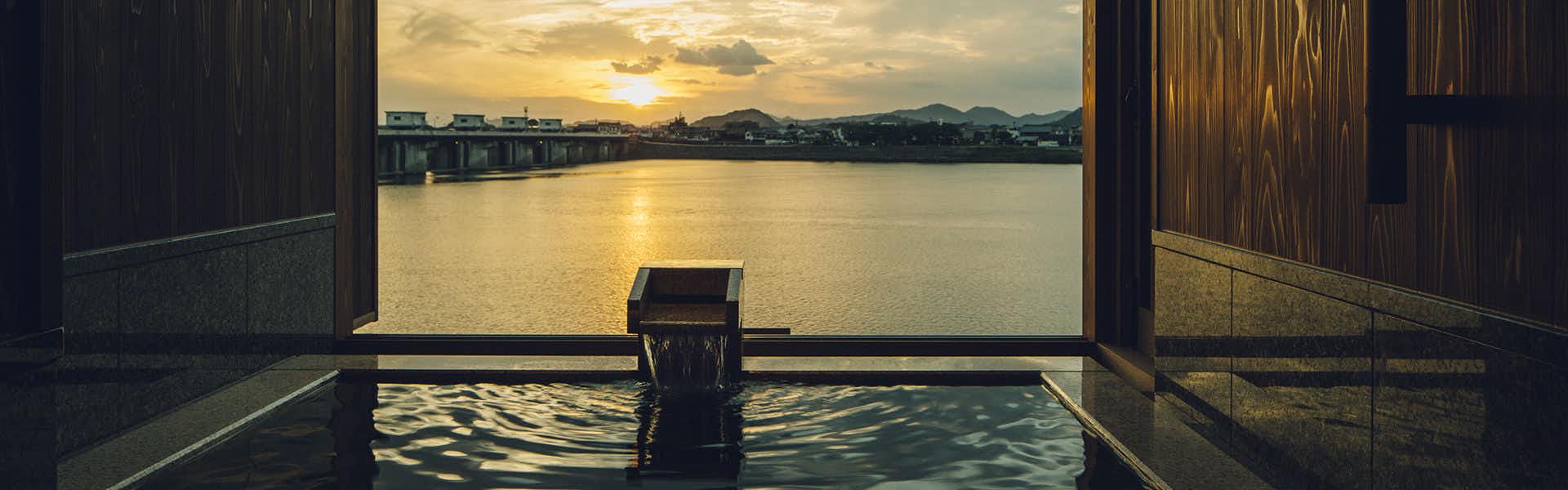 Akariya Geihanro Interior bath with large wooden-framed window framing sunset light on water and distant mountains.