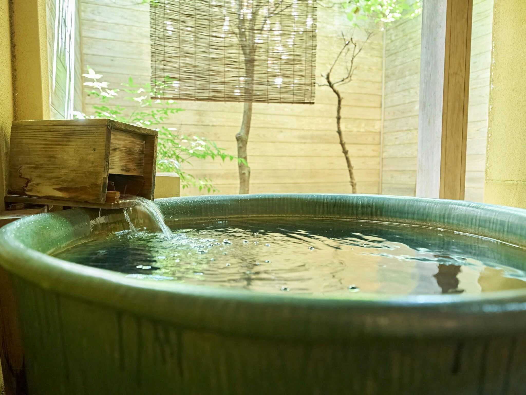 Ohamaso Round green ceramic bath with a wooden water spout beside a bamboo blind, pale wall, and small courtyard tree