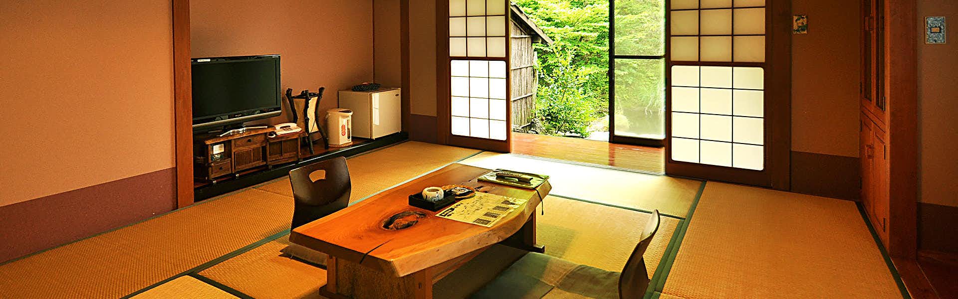 Genryu no Yado Hozantei Brown walls, natural wood flooring, black chair, low wooden table, large gridded window panes, exterior vegetation visible.