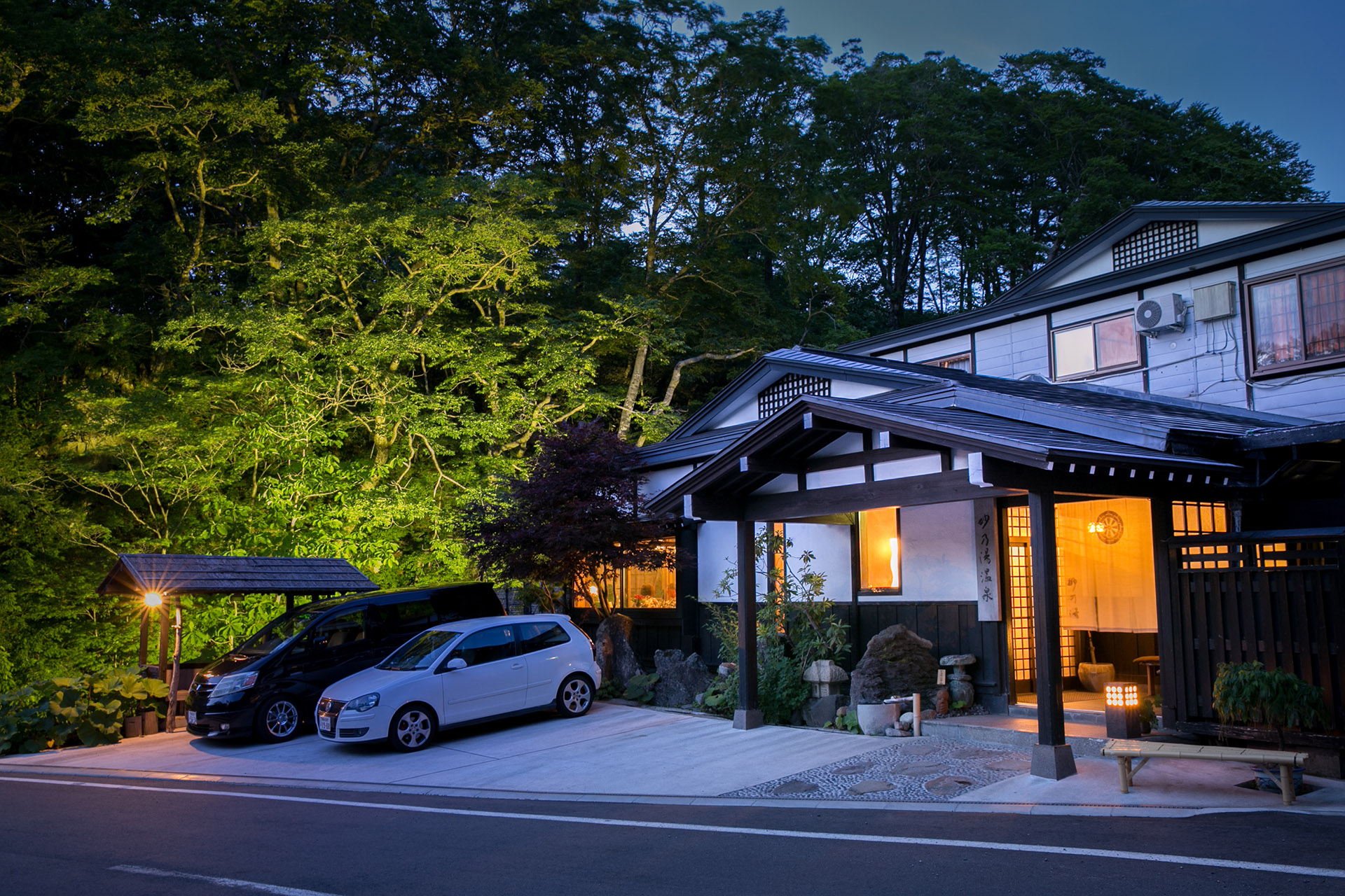 A warmly lit timber entrance sits at the forest edge, with traditional rooflines, stone landscaping, and a covered drop-off.
