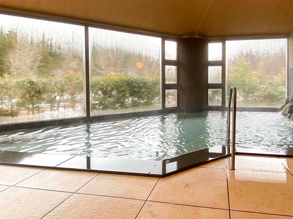 Maruoka Onsen Takekurabe with a broad indoor bath beside windows overlooking winter trees.