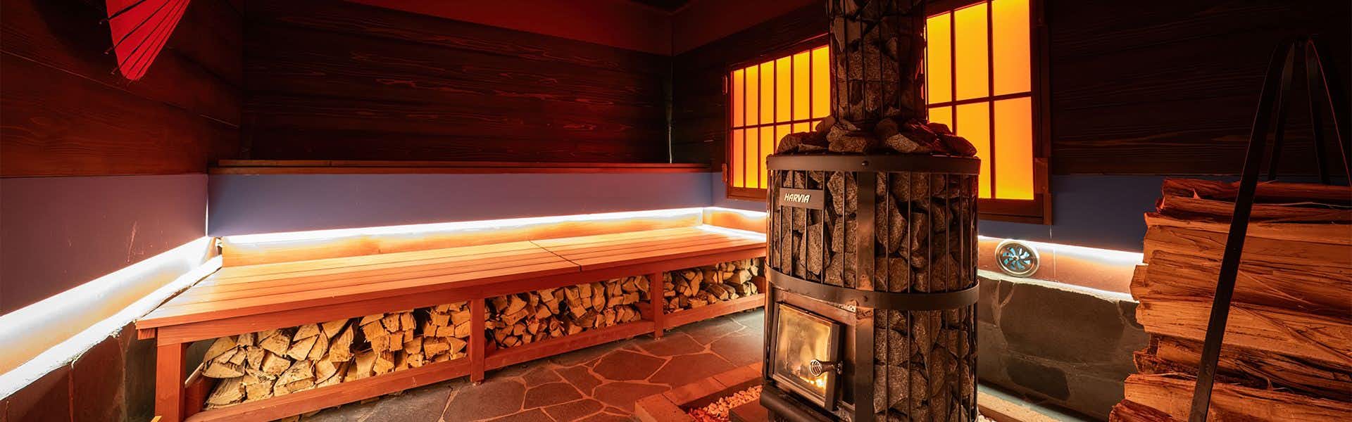 Mountain Inn Kan no Jigoku Ryokan Red-lit sauna with long wooden bench, stacked firewood, central metal stove filled with stones, and glowing windows