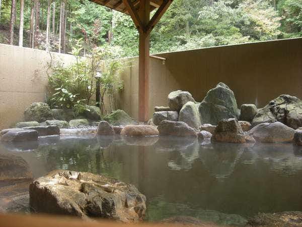 Sugatani Onsen Rock-lined outdoor bath beneath a timber roof with large boulders, garden shrubs, and forest beyond
