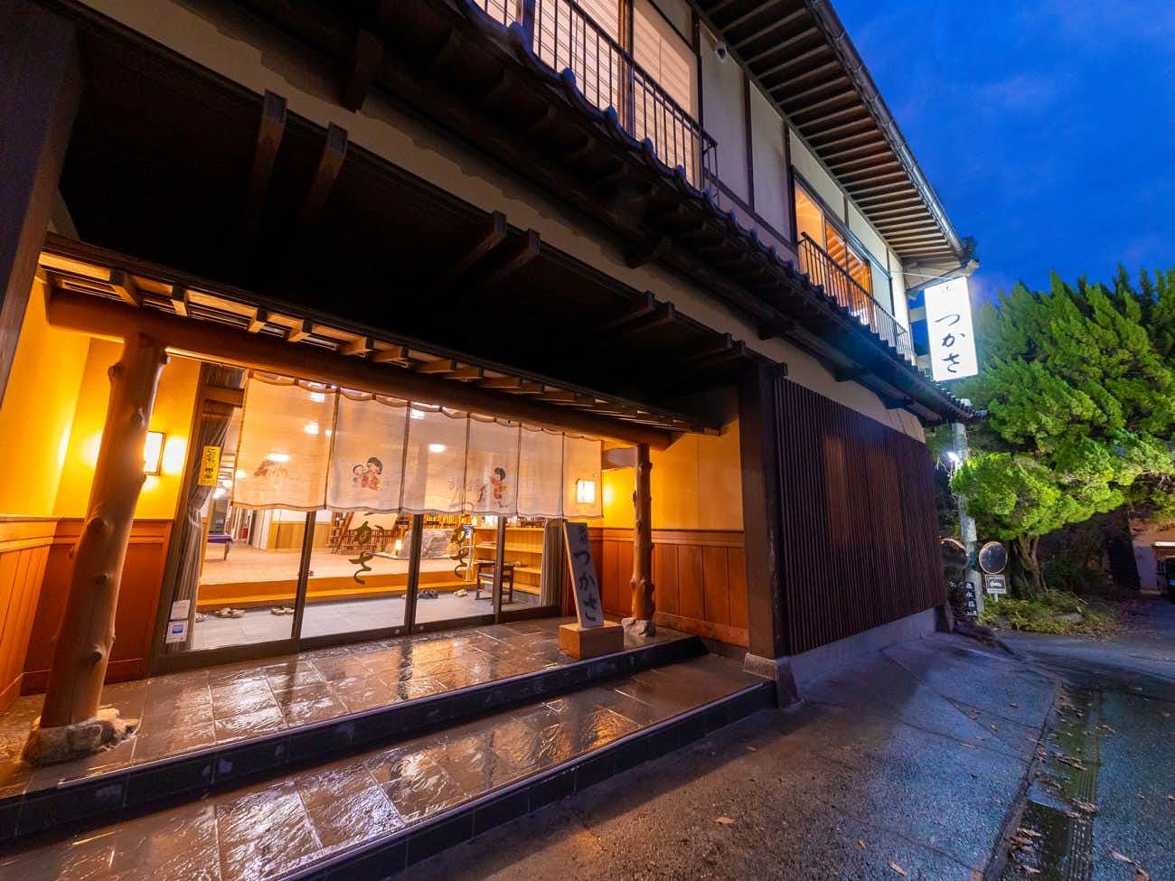 Furuyu Onsen Tsukasa Ryokan Multi-section wooden building facade with illuminated windows, horizontal wooden railings on upper level, wooden siding, blue sky above roofline, green foliage at corner, wet street surface with reflections