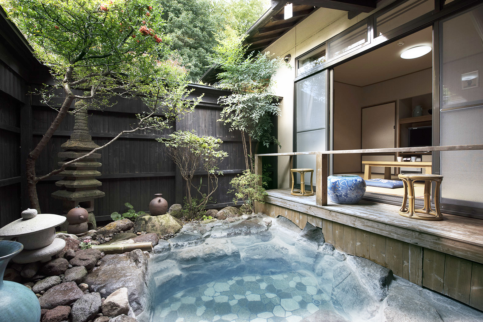 A stone-lined outdoor bath fills a courtyard beside a wooden veranda and lush garden.