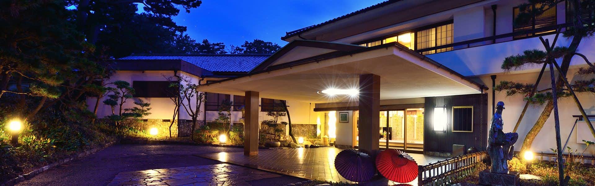 Hagoromo Hotel Building exterior with warm landscape lighting, purple courtyard surface, red objects, and garden plantings at dusk.
