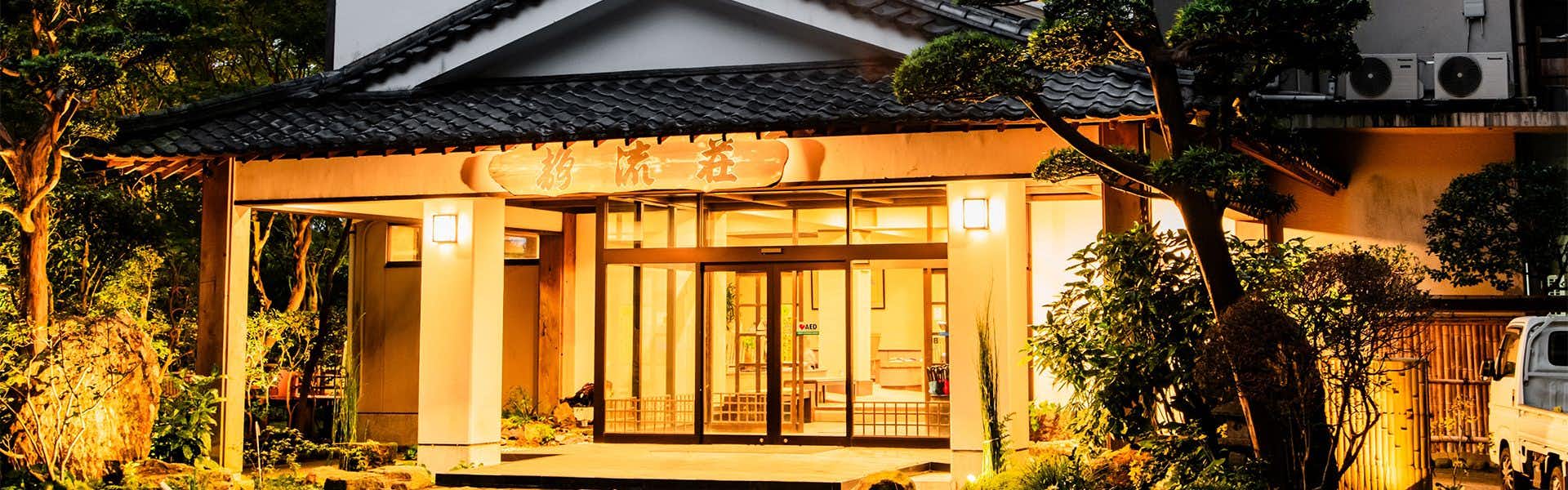Kirishima Momiji Valley Seiryuso Building exterior with amber ground lighting, wooden vertical cladding, windows, and tree foliage framing one side at dusk