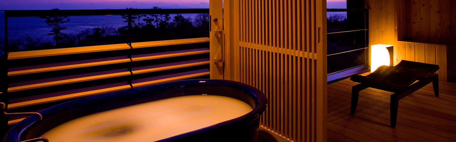 Himakajima Island Villa Yuzuki Oval bath under warm amber lighting beside wooden slat screens, low chair, lantern, and twilight sea view