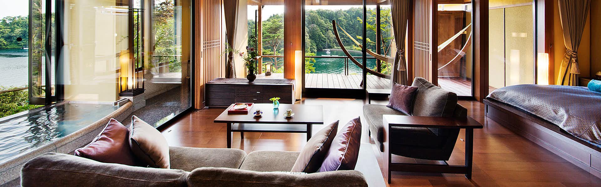 Noto Tsukumowan Hyakurakuso Interior living space with wood floors, neutral upholstered furniture, wooden columns, and windows overlooking forested landscape.
