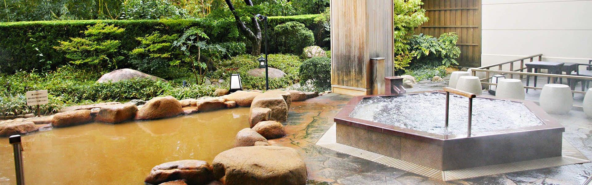 Hyoe Koyokaku Outdoor bath facility with tan-colored water, large boulders, metal railings, white stone edge, and dense green vegetation.
