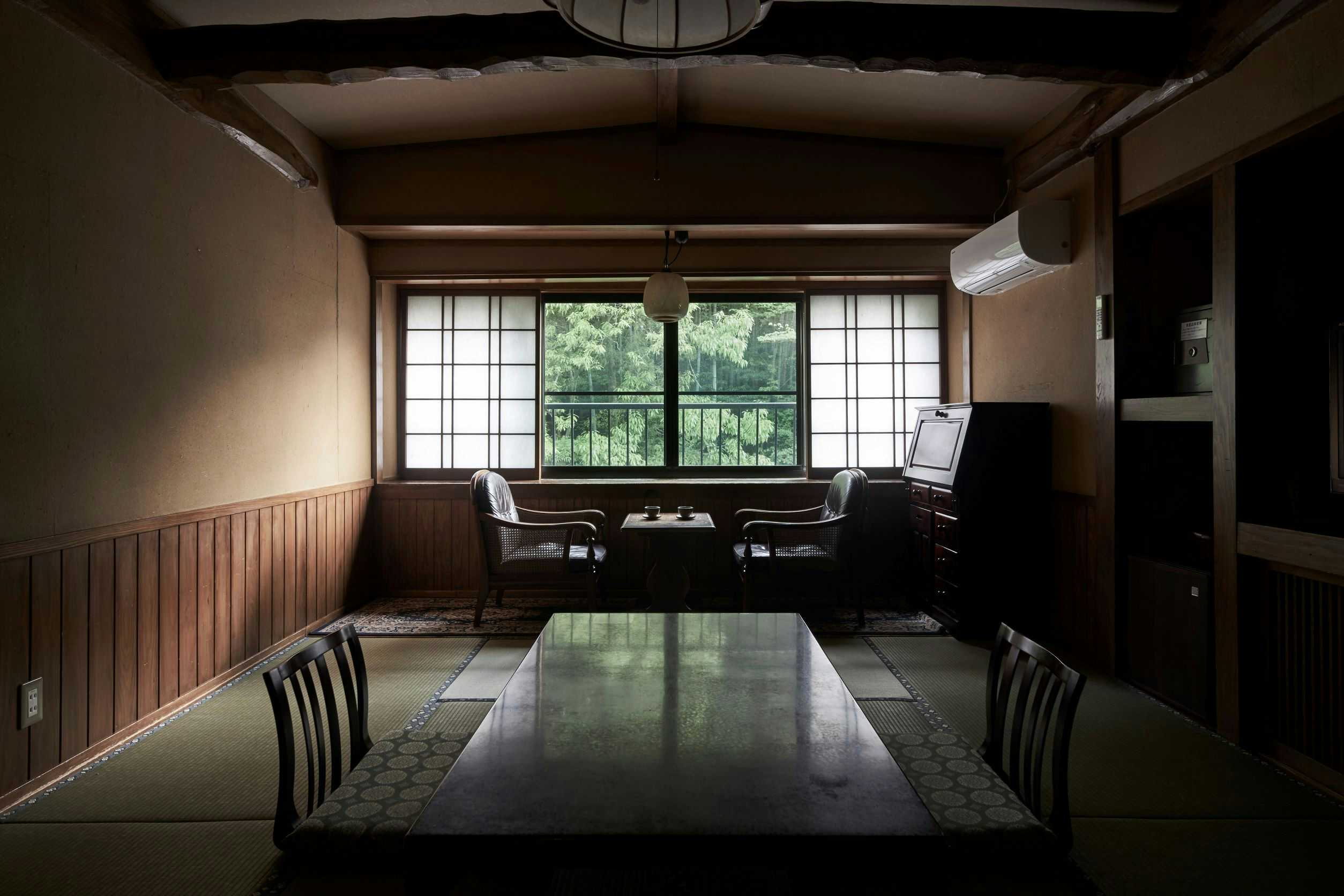Fumoto Ryokan dining space with long wooden table, dark wood ceiling beams, grid windows overlooking foliage, traditional seating, wood railings.