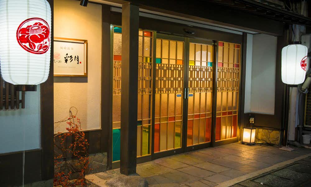 Hanakoji Saigetsu Illuminated entrance with white lanterns, gold-framed glass doors, dark timber posts, and a stone walkway