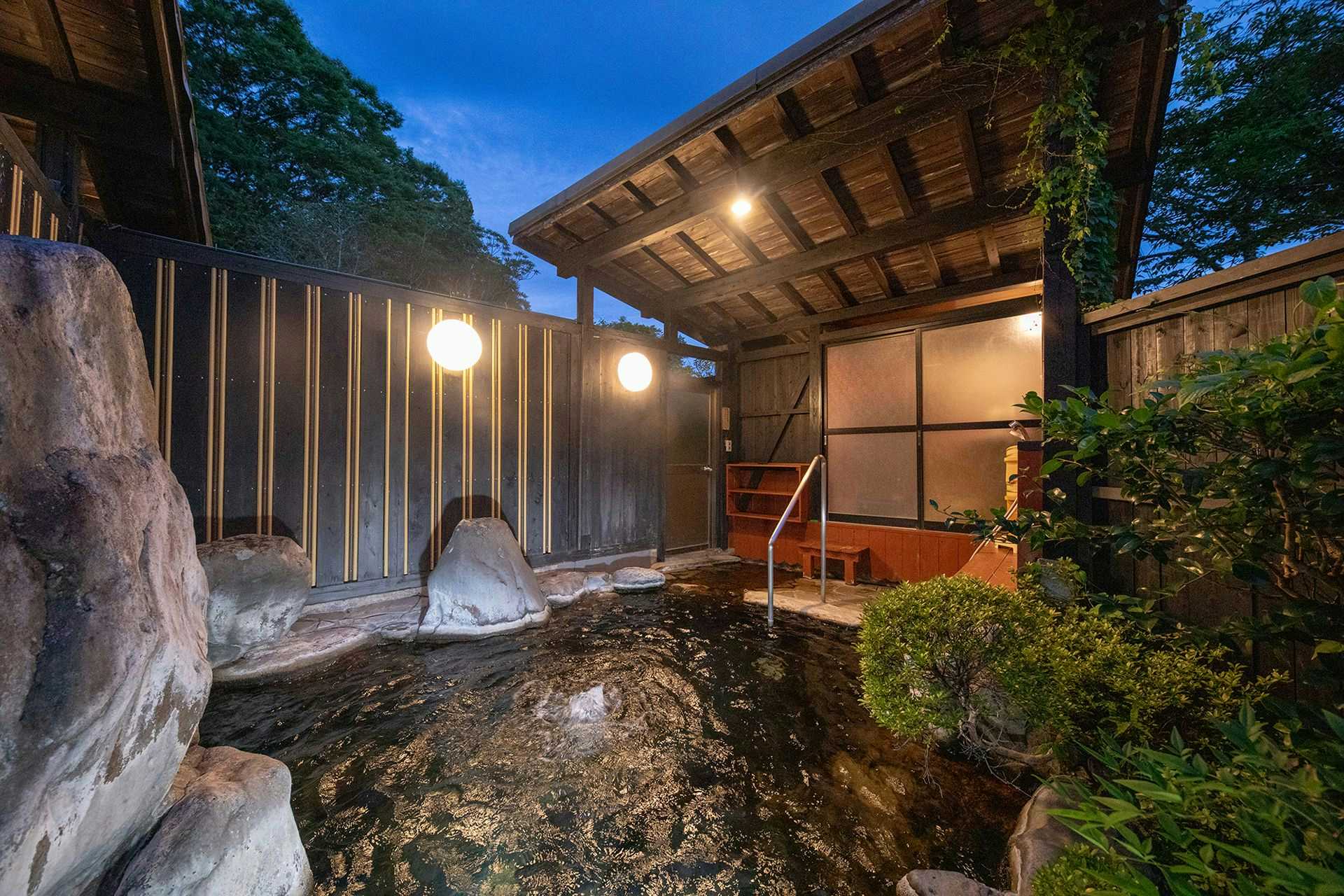 Yufuin Onsen Villa Konjakuan with this scene, where a rock-lined private open-air bath sits beneath a timber roof and garden trees.
