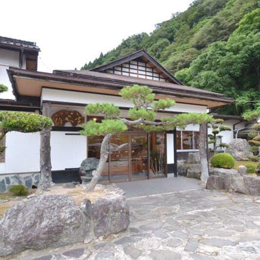 Yumura Onsen Hakuntei with a stone-paved entrance framed by clipped pines and green mountains.
