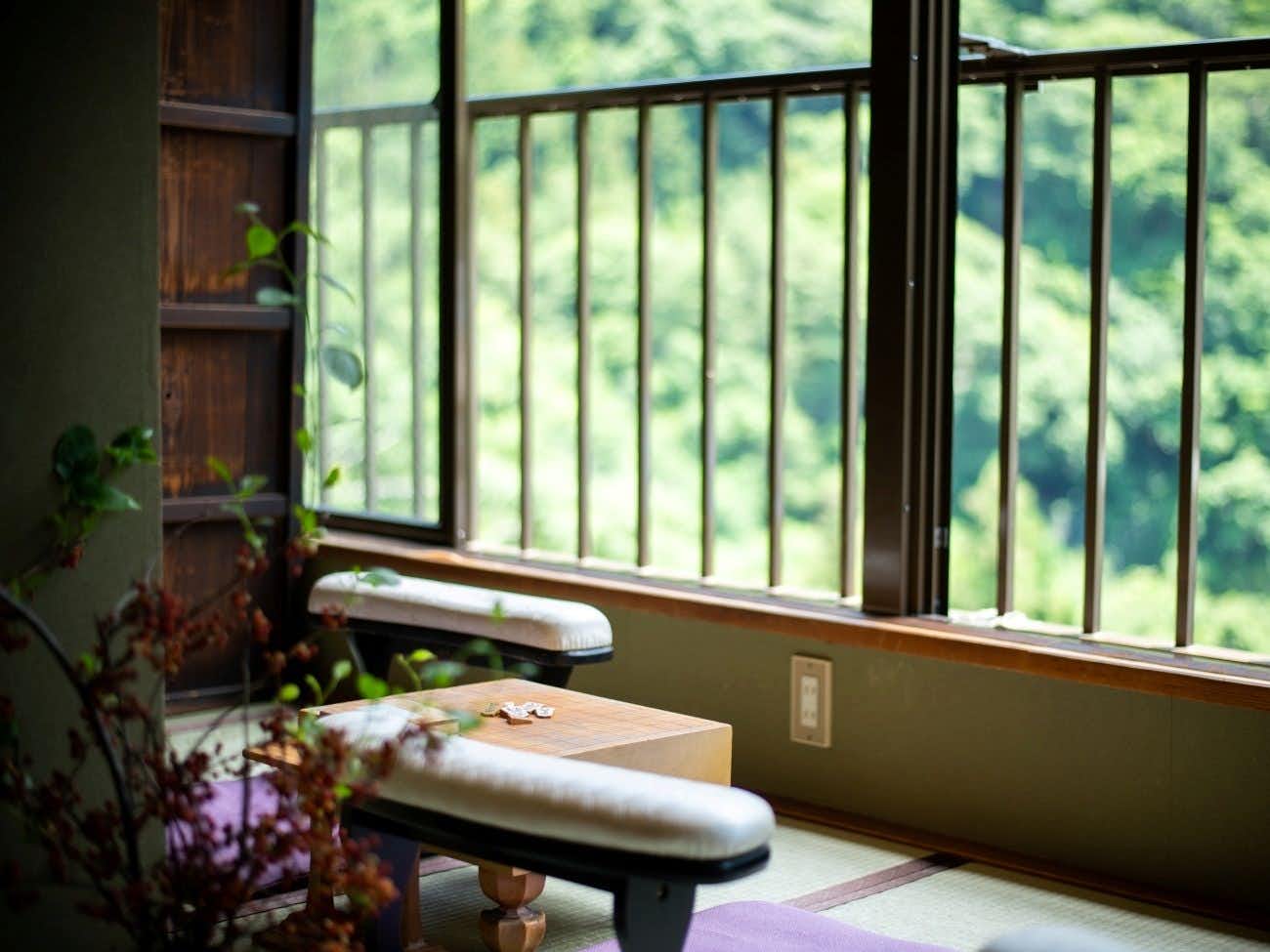 Gorokaku Window-side seating nook with a low wooden table, upholstered stools, flowers, and soft green hills beyond