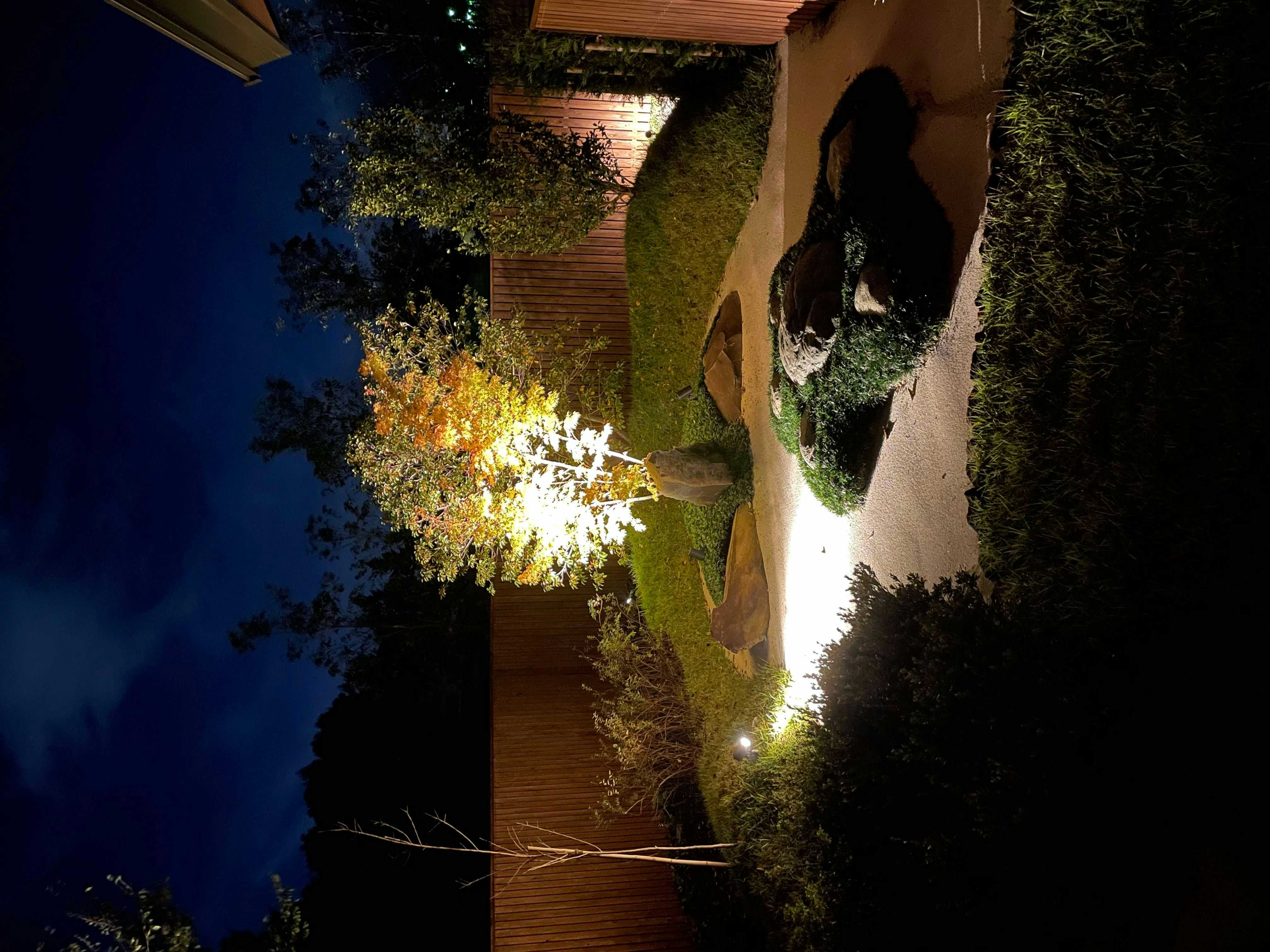 Hakkai konohanakan Illuminated night garden with pale gravel, grass, boulders, shrubs, trees, and wooden boundary walls