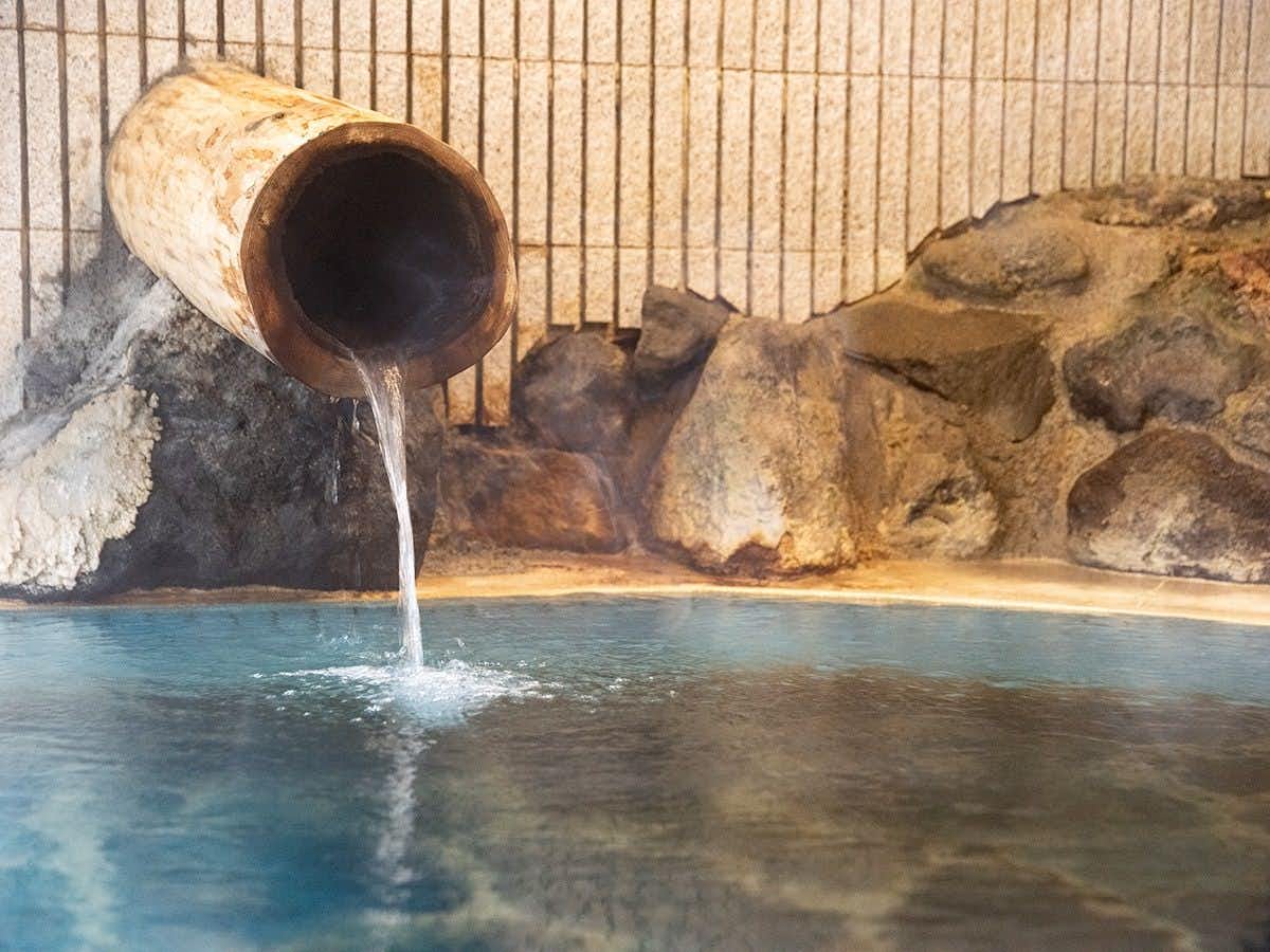 Futamata Onsen Kashiwaya Ryokan Metal pipe discharging into pool bordered by large rocks with bamboo slat fence visible in background
