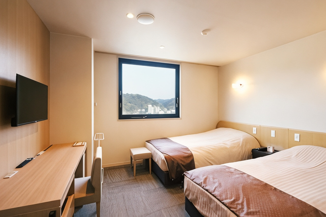 A bright room contains two beds, a long desk with chairs, a wall mounted television, and a window overlooking hills.