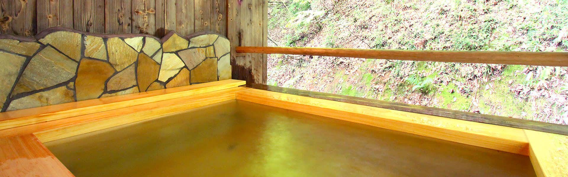 Yanagiya Ryokan Bath interior with yellow-tinted water, large window frame revealing forest trees and vegetation, horizontal wooden beam crossing