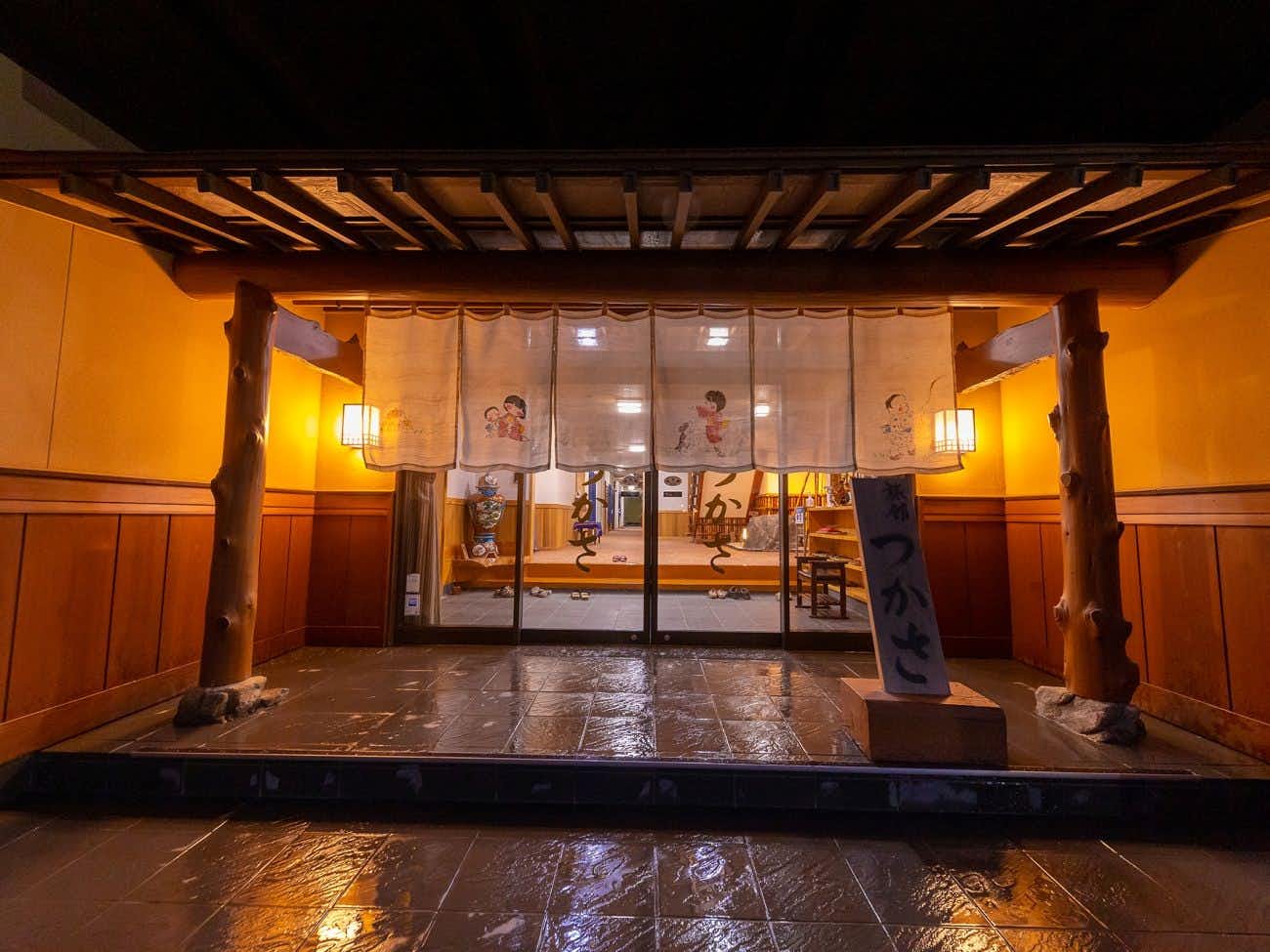 Furuyu Onsen Tsukasa Ryokan Wooden entrance structure with deep overhanging eave, exposed crossbeams, illuminated display windows with interior figures visible, vertical wooden support columns, vertical signage post on left, wet ground below