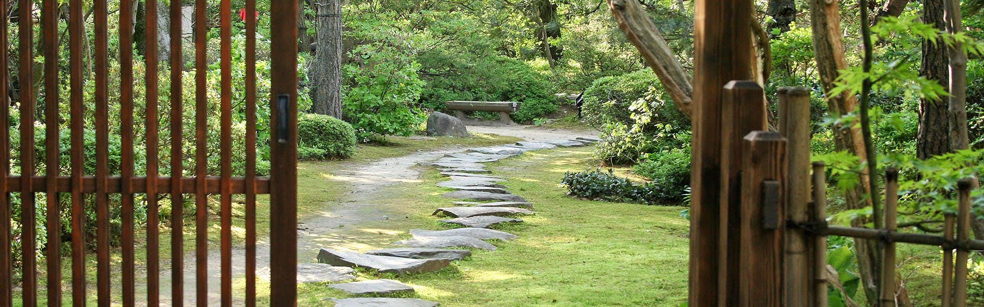 Shikisai Ichiriki Stone rectangular stepping stones set through grass pathway beneath overhead tree canopy with surrounding green plantings.