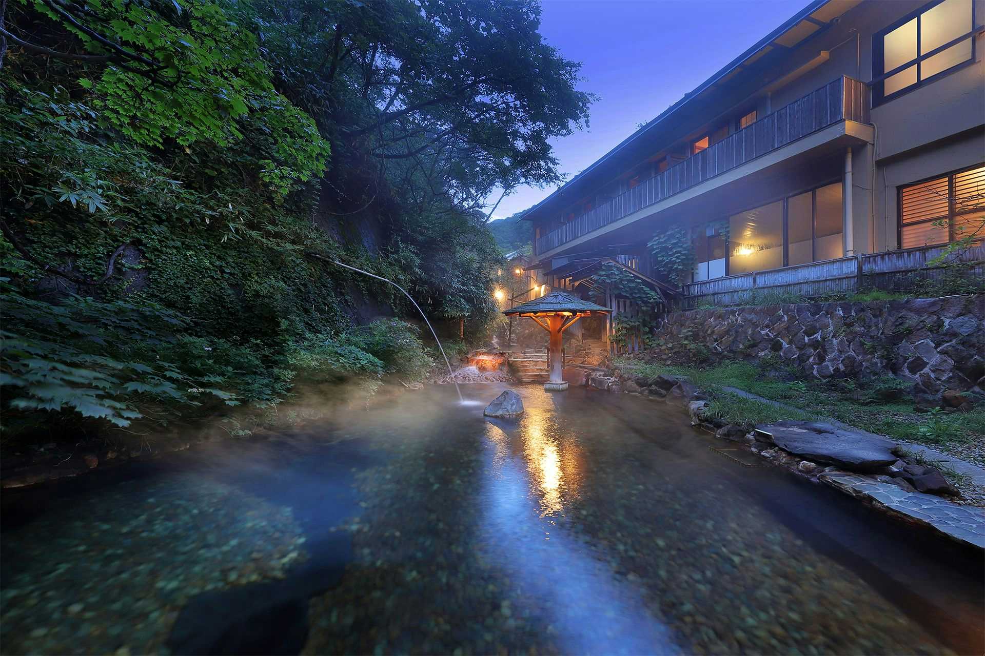 Oomaru Onsen Ryokan with the riverside building glowing beside a steaming outdoor bath.