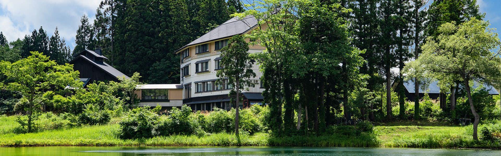 Oshimizu no Yu Tsutaya Cream-colored lodge with a dark pitched roof beside a still pond, surrounded by tall trees and dense greenery.