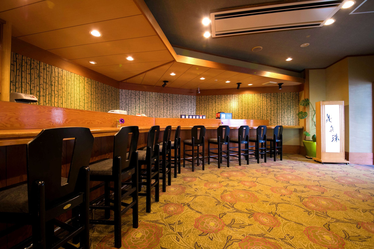 A long wooden counter is lined with tall black chairs against a bamboo-patterned wall.