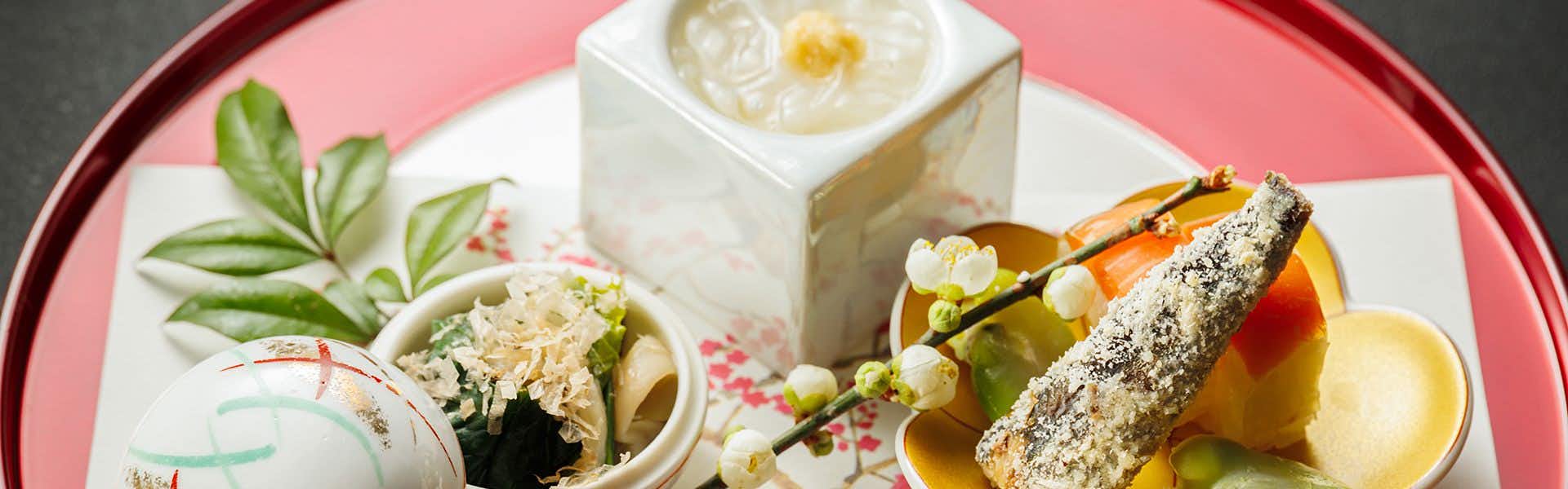 Ryotei Jyusouya Sliced items on coral-pink plate with white ceramic vessels, green herb sprigs, and pink flowers arranged beside
