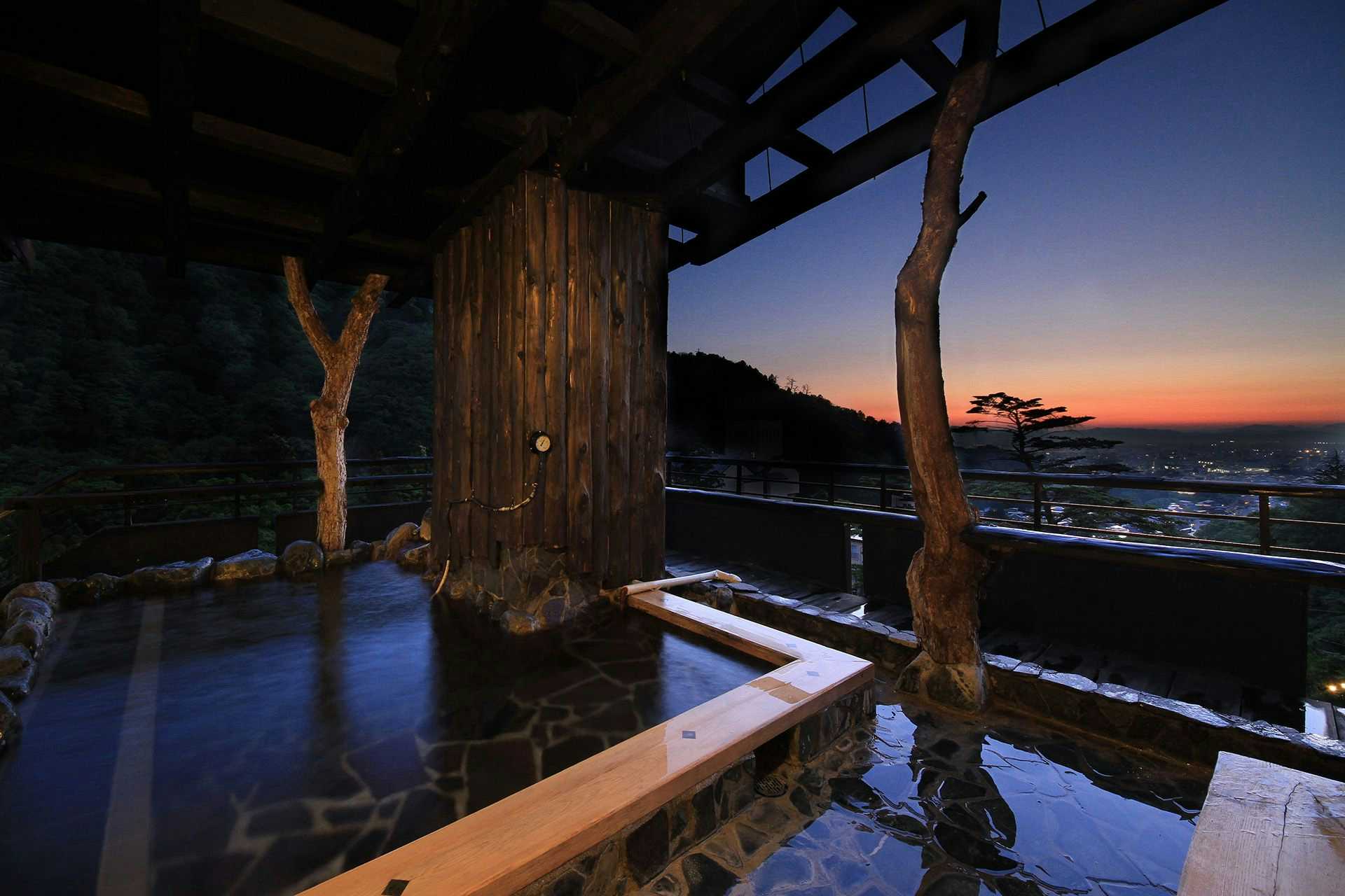 Kutsurogijuku Shintaki Roofed outdoor stone bath at twilight beneath timber beams with a broad town and hillside outlook