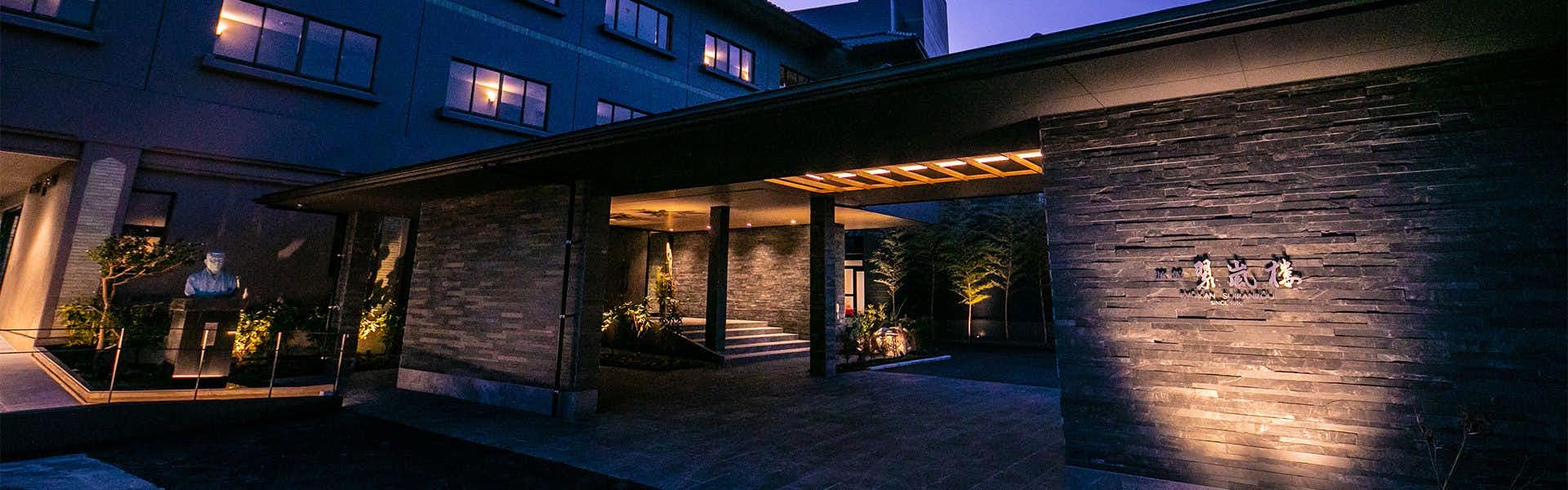 Hitoyoshi Onsen Ryokan Suiranro with a dark stone entrance, covered driveway, lit sign, and evening sky