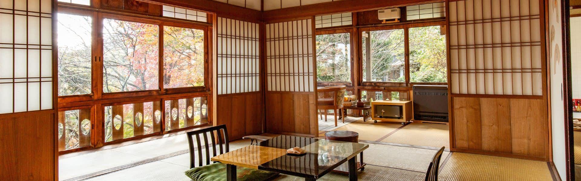 Senzairo Dark wooden lattice screens, low wooden tables, cushioned seating, windows with exterior foliage visible, light-colored flooring.