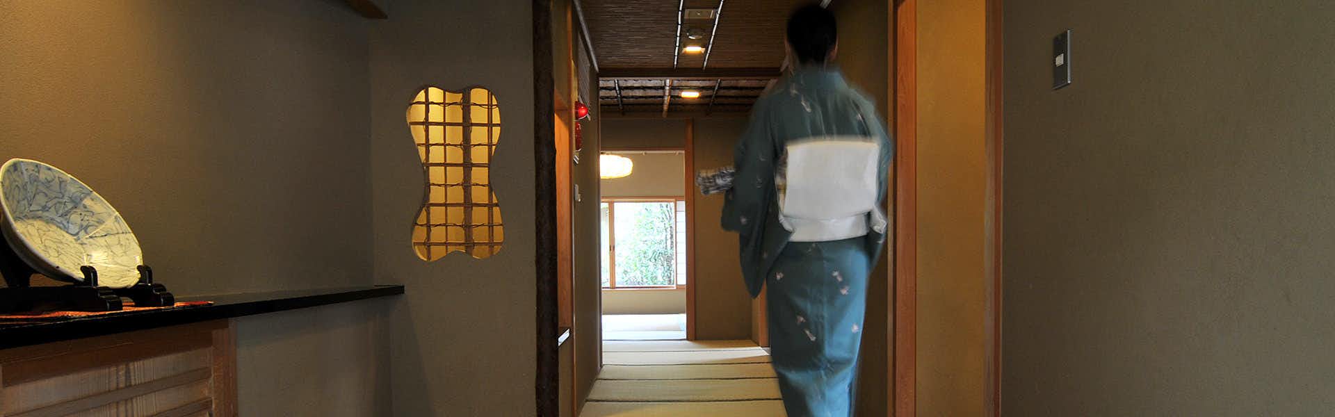 Atami Sekitei hallway with person wearing dark jacket and white towel across back, wooden latticed panel doors, wood ceiling beams visible, wood flooring, doorway showing outdoor light