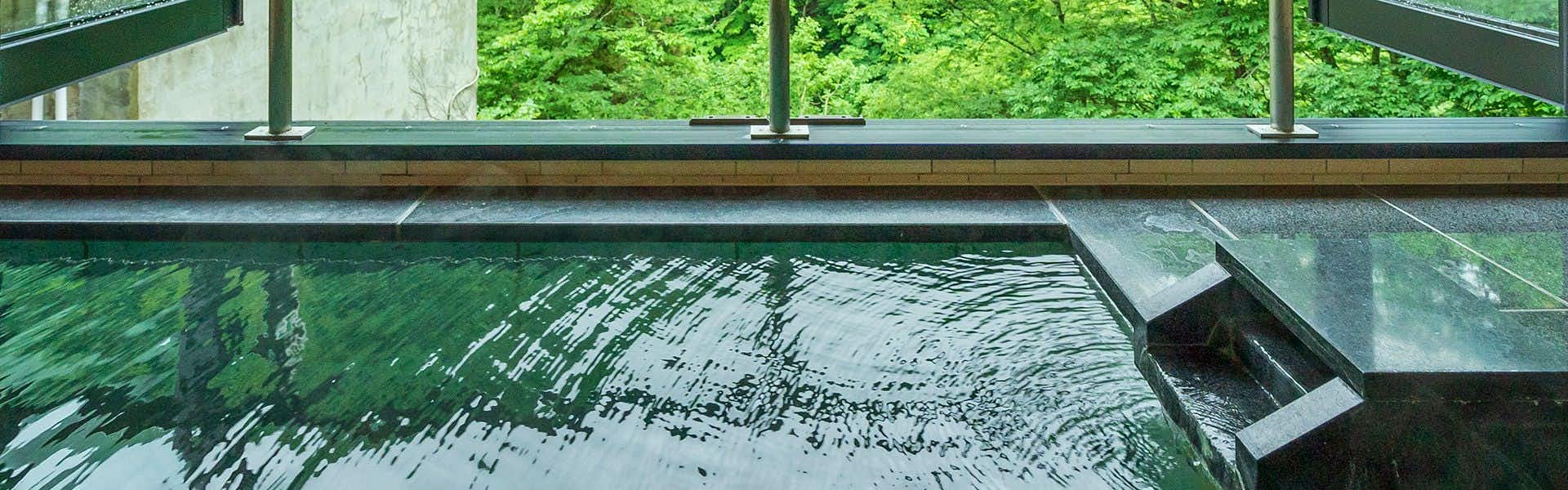 Shinshu Yamada Onsen Yamadakan Indoor bath with blue-tinted water, large window viewing green forested landscape, wooden frame and stone bath edge.