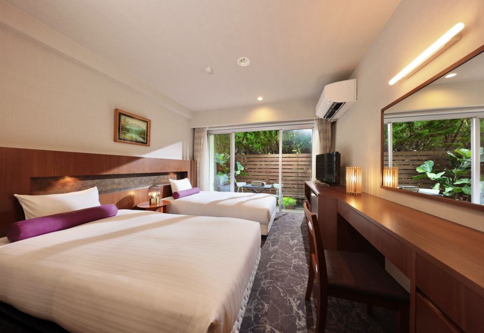 A twin bedroom with a long desk, illuminated mirror, and glass door to greenery.