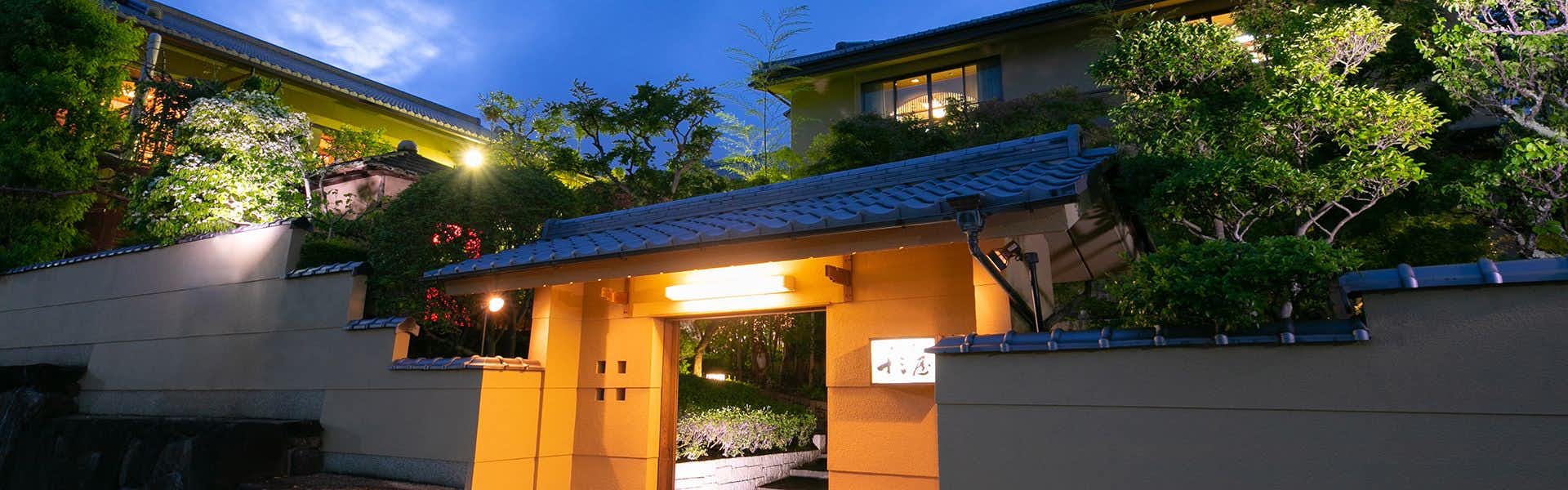 Ryotei Jyusouya Exterior entrance with yellow-lit wooden doors, blue-tiled roof, mounted lantern, and mature trees surrounding building