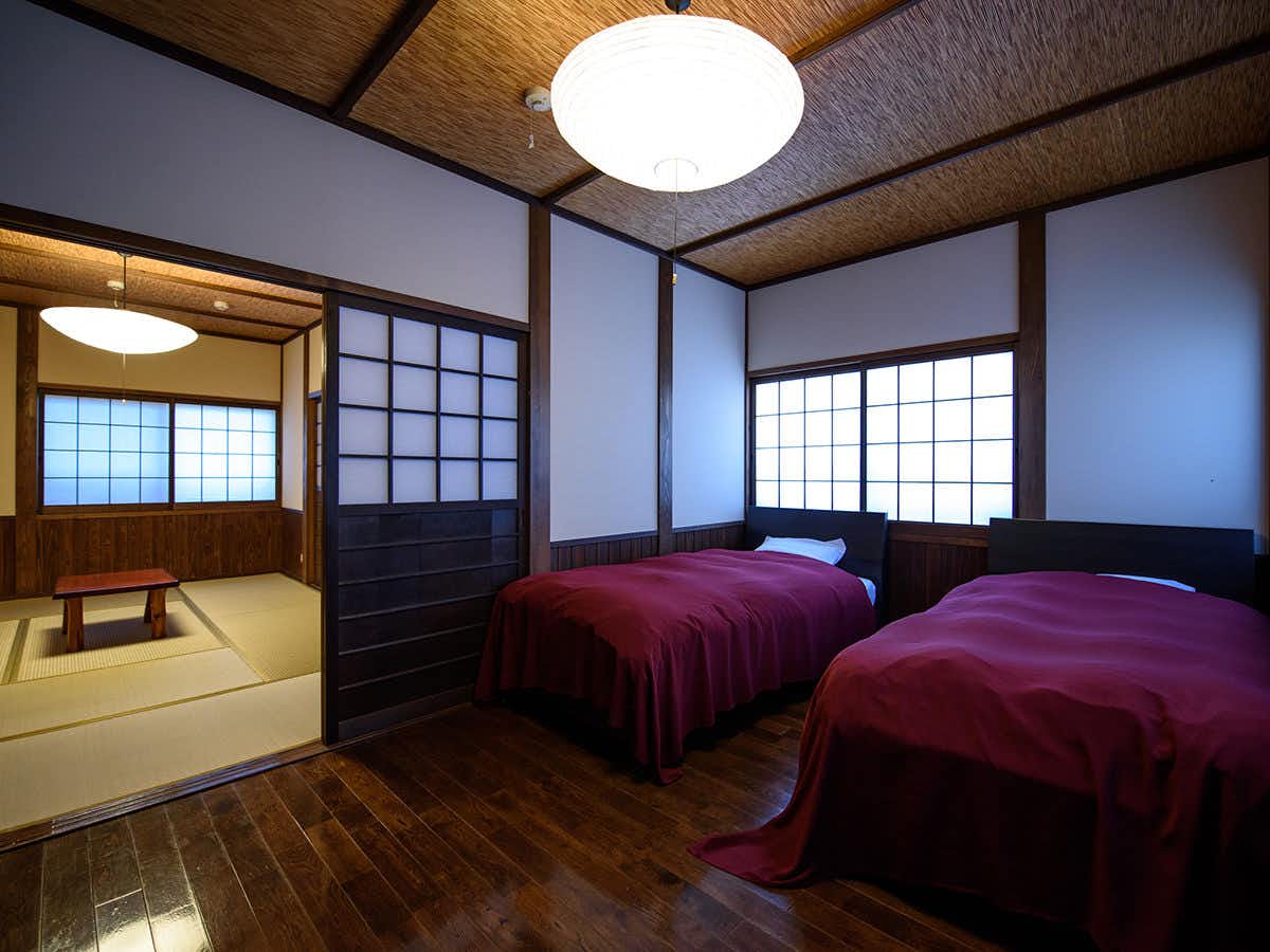 Yunohira Onsen Ryokan Kamiyanagiya Bedroom with two burgundy beds, tatami flooring, dark wooden storage, gridded shoji screens, and wooden beamed ceiling.