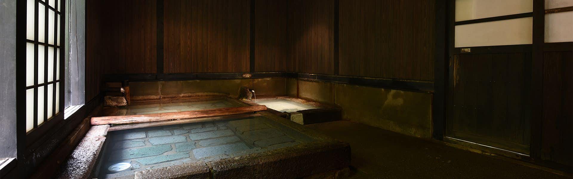 Mountain Inn Kan no Jigoku Ryokan Dim wood-paneled bathing room with rectangular stone-lined pool, water spouts, textured floor, and tall window