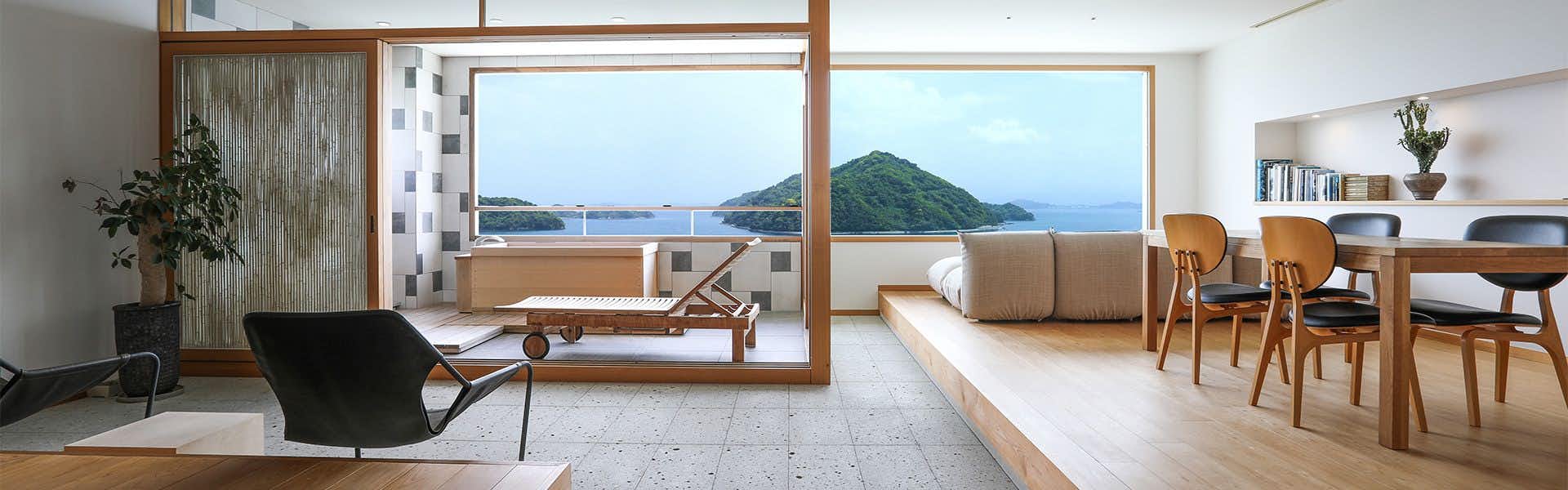Kinoe Onsen Hotel Seifukan Bedroom with large wooden-frame window, white bedding, wooden lounger, ocean and forested island visible through glazing