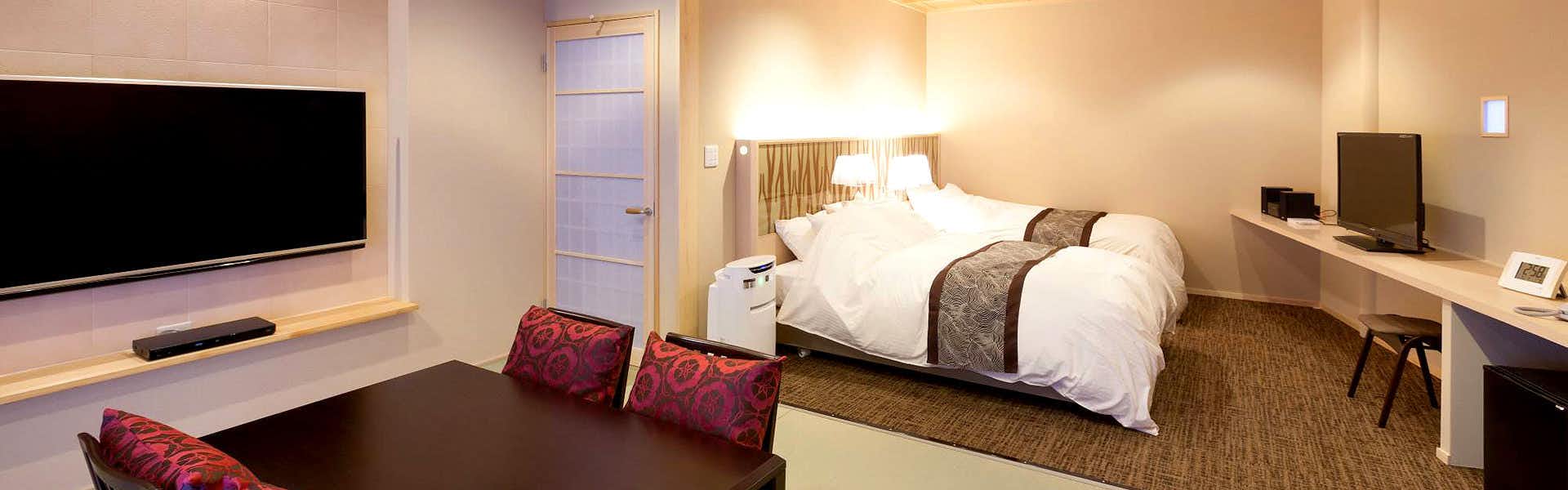 Matsuba Onsen Taki no Yu Contemporary bedroom with two white beds, patterned runners, large wall-mounted television, dark table, armchairs, desk, and soft lighting