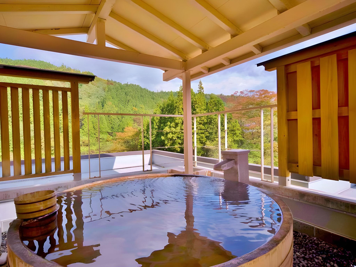 A round outdoor wooden tub beneath a roof overlooking green woodland.