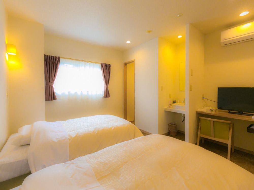 Sokai no Yado Narai no Kaze Bedroom interior with two white beds, yellow walls, curtained window, desk area with screen, warm lighting.