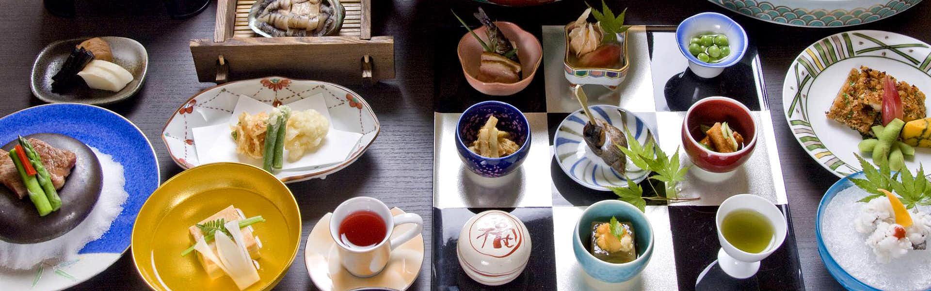 Matsuba Onsen Taki no Yu Assorted small dishes, bowls, cups, and plates arranged across a dark dining table with colorful ceramic tableware